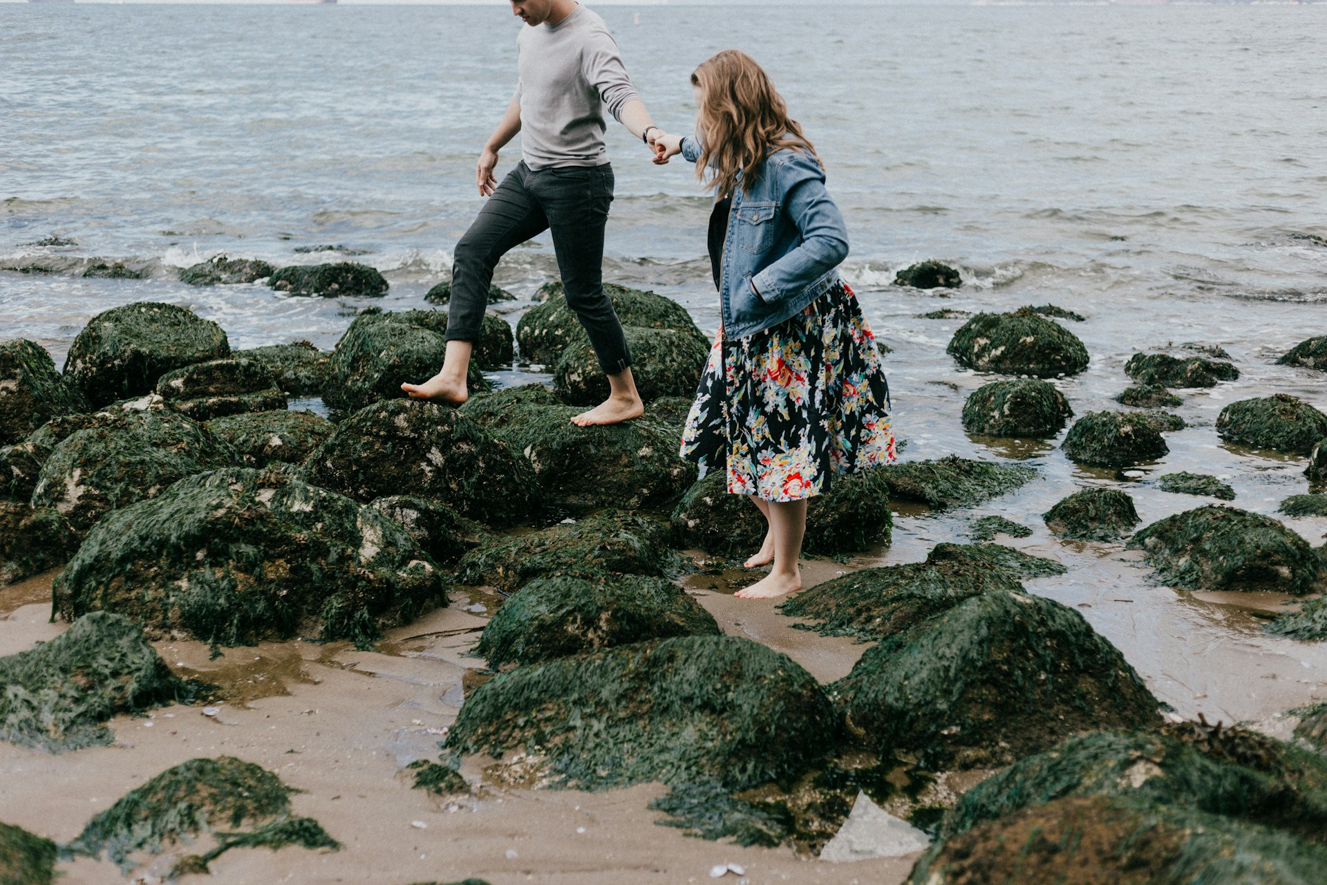 A man and woman holding hands stepping over rocks near the sea